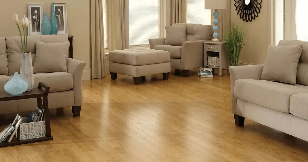 A living room with professionally installed bamboo flooring, beige sofas, a matching ottoman, a side table with a blue lamp, vases, framed pictures, and large windows with blinds and curtains in San Diego, CA