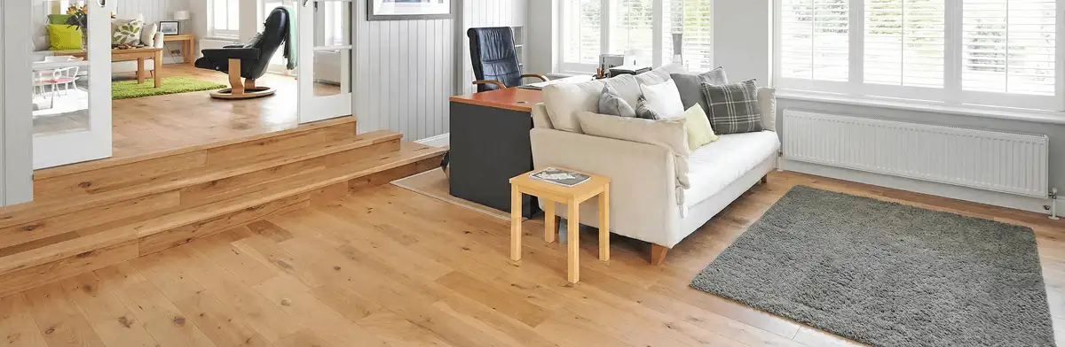 A modern living room with professionally installed laminate flooring, a beige sofa, a small side table, a gray rug, and large windows with white shutters in San Diego, CA